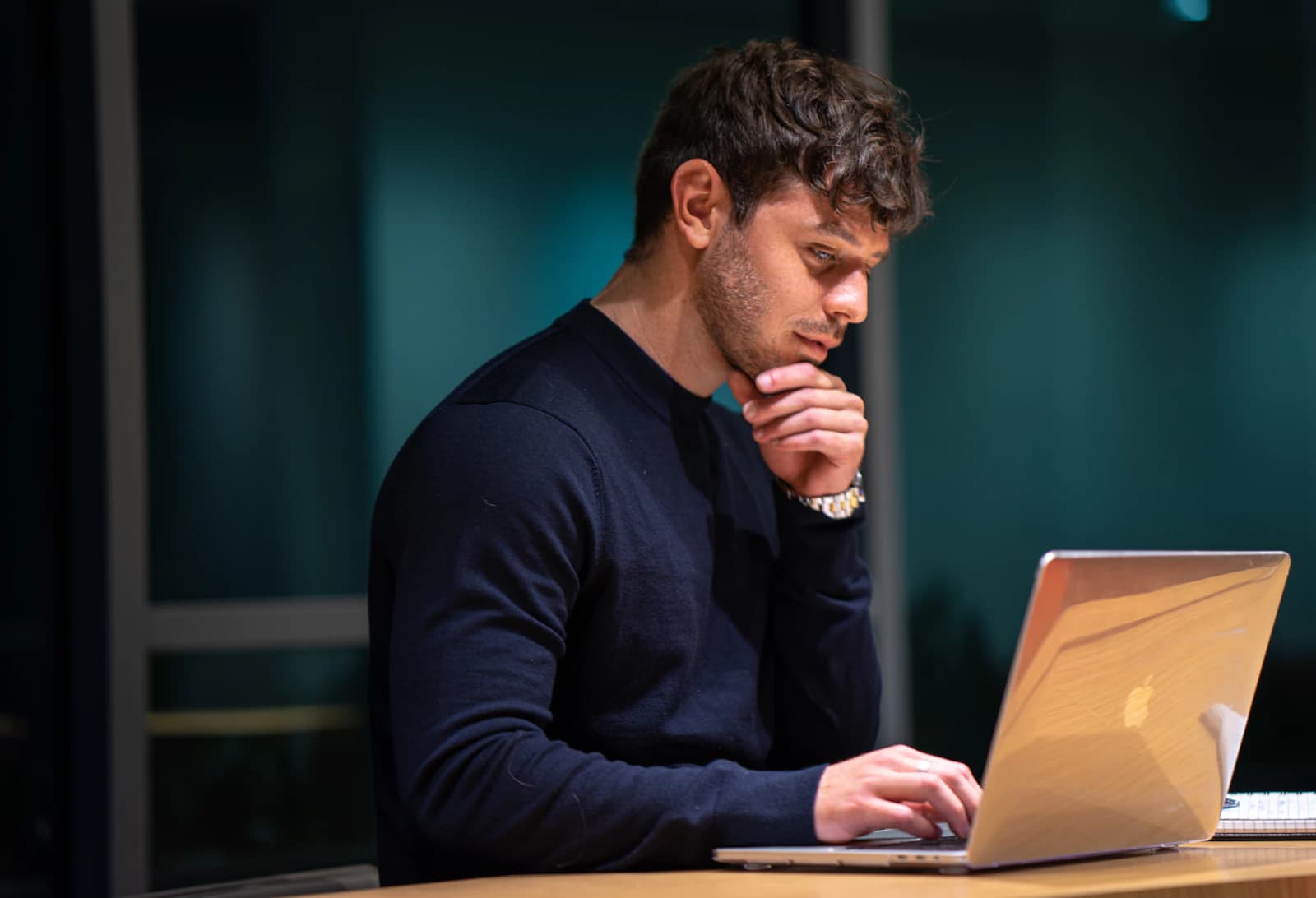 A man in a dark sweater working at a laptop with his hand resting on his chin
